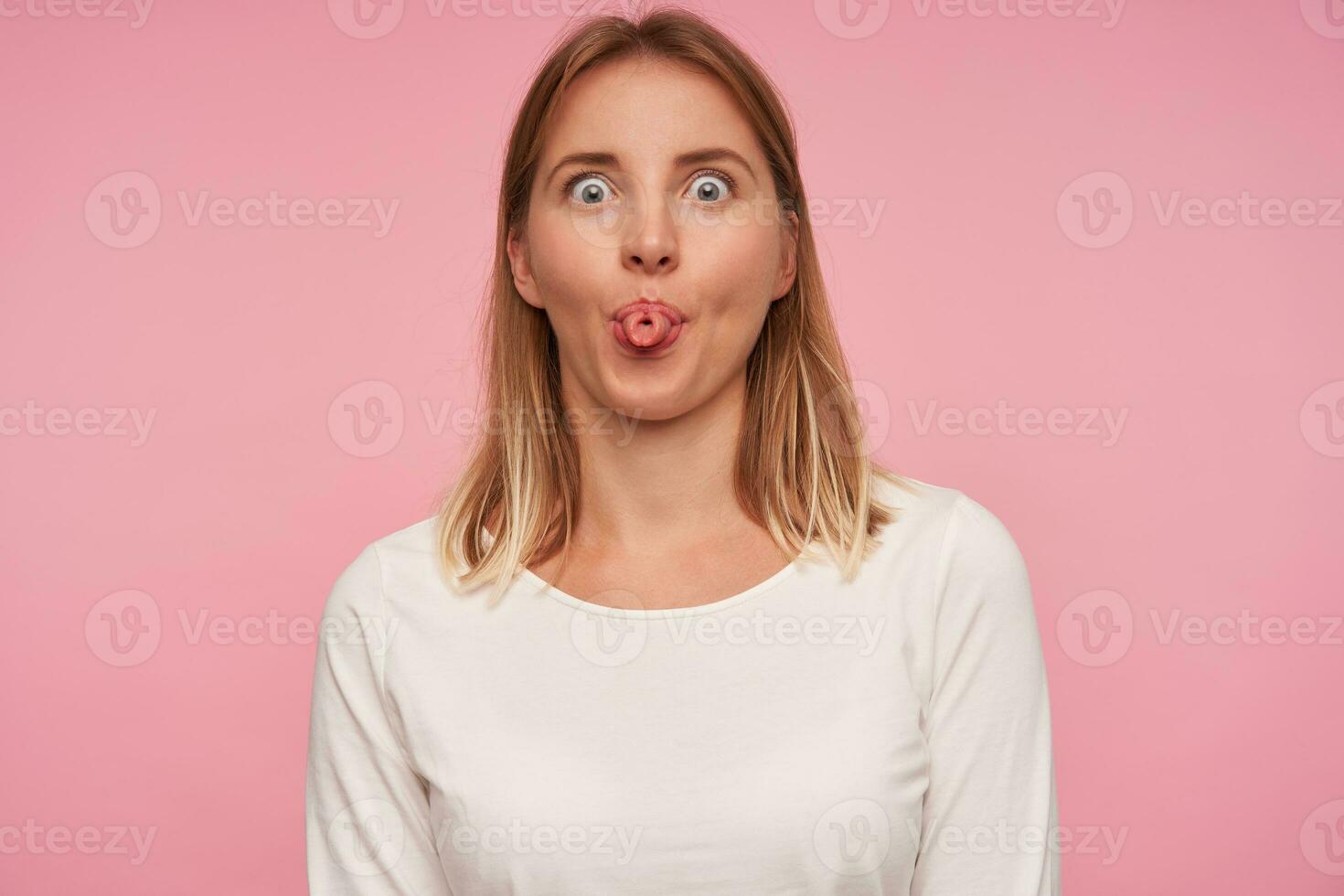 Positive pretty young blonde female with casual hairstyle making fun over pink background, rounding eyes and sticking out folded tongue, wearing white blouse photo