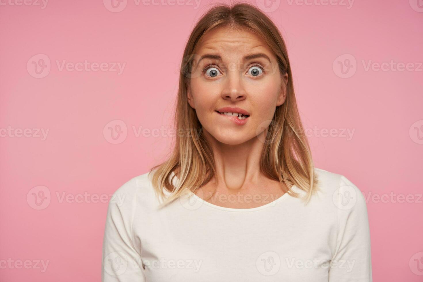 Shocked attractive young blonde woman biting underlip and looking at camera with wide eyes opened, raising eyebrows and wrinkling forehead over pink background photo
