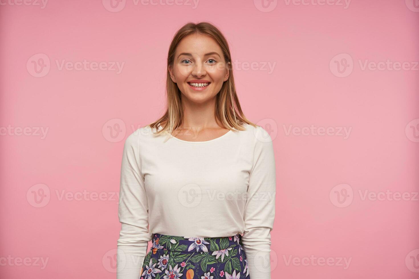 Indoor photo of charming young blonde woman posing over pink background with wide sincere smile, keeping hands along body, demonstrating her white perfect teeth