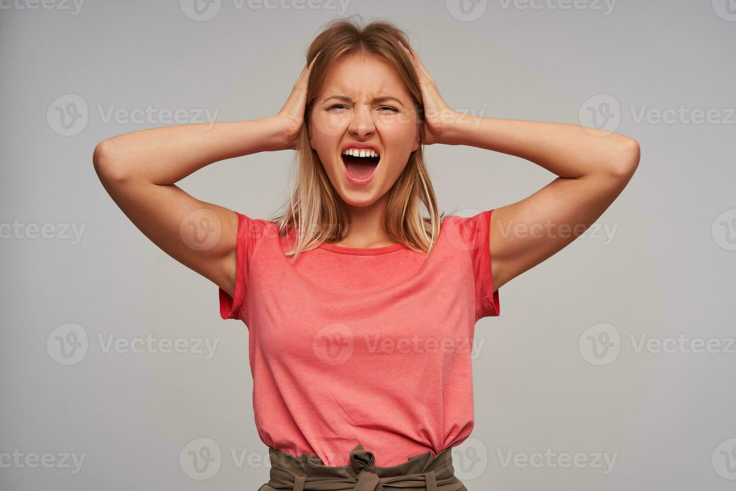 Stressed young pretty blonde female with casual hairstyle holding her head with raised hands, frowning her face with wide mouth opened while looking at camera, isolated over white background photo