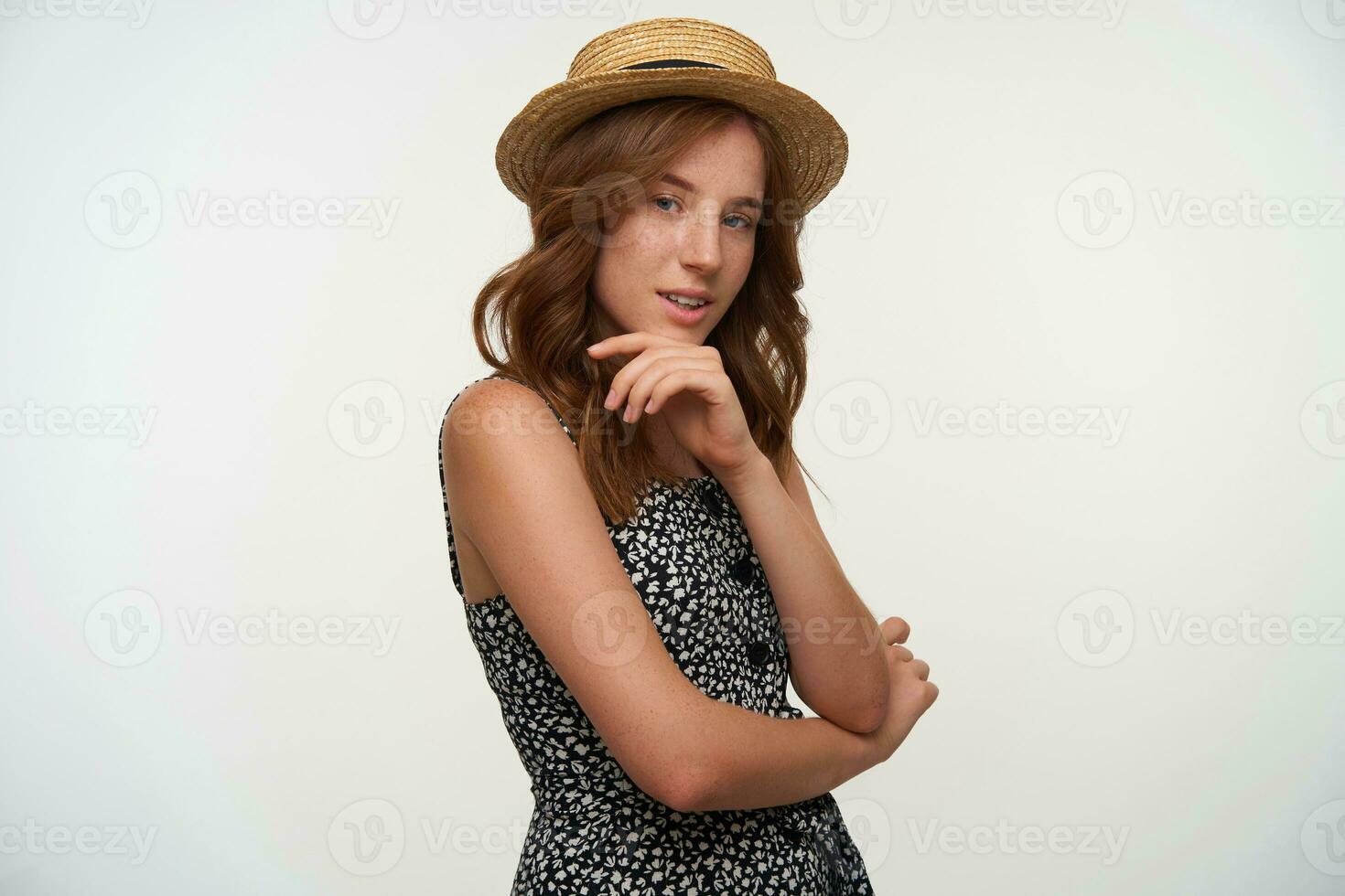 Studio shot of beautiful young female with red wavy hair looking to camera thoughtfully and keeping hand on her chin, posing over white background in romantic dress and straw hat photo