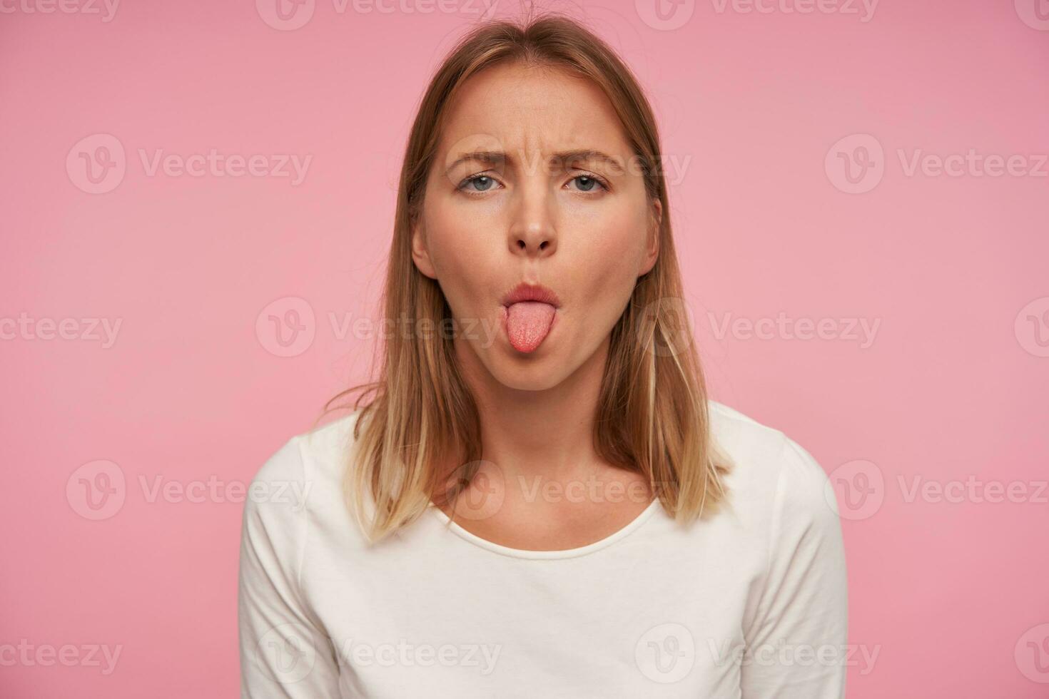 Portrait of beautiful blonde female with casual hairstyle frowning eyebrowns and showing her tongue while posing over pink background, wearing white blouse photo