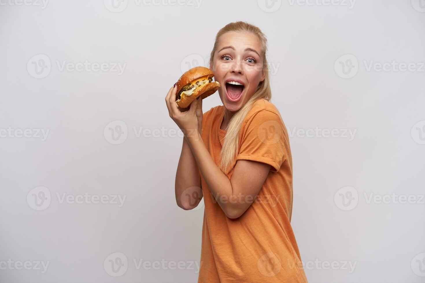 Studio photo of young blue-eyed excited blonde female dressed in orange t-shirt holding big burger in raised hands and looking at camera with wide eyes and mouth opened, isolated over white background