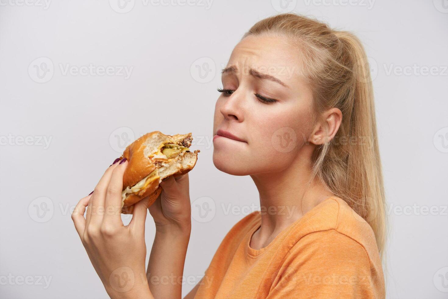 retrato de joven encantador largo peludo rubia hembra con cola de caballo peinado acuerdo hamburguesa en elevado manos y mirando insaciablemente en él, mordiendo debajo del labio y ceñudo Cejas terminado blanco antecedentes foto