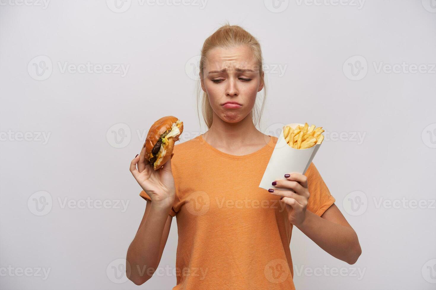 trastornado joven bonito rubia mujer en naranja camiseta acuerdo insalubre comida en su manos y mirando tristemente a él, ceñudo Cejas y retortijón su boca mientras posando terminado blanco antecedentes foto