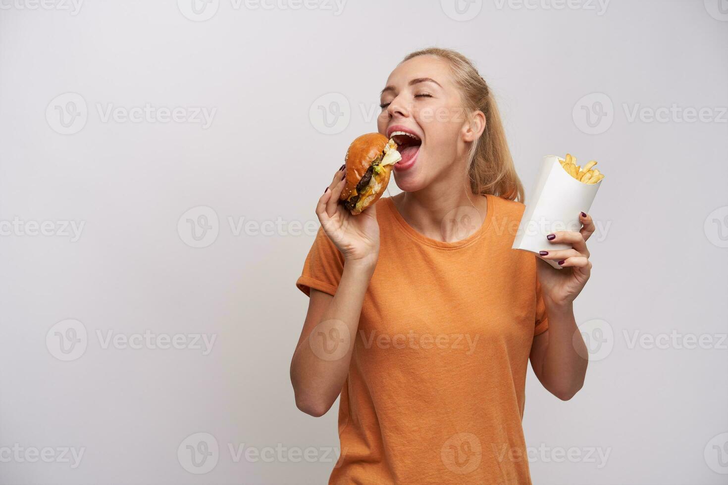 Pleasant looking positive young blonde lady with ponytail hairstyle keeping eyes closed while enjoying her tasty burger, standing over white background in casual clothes photo