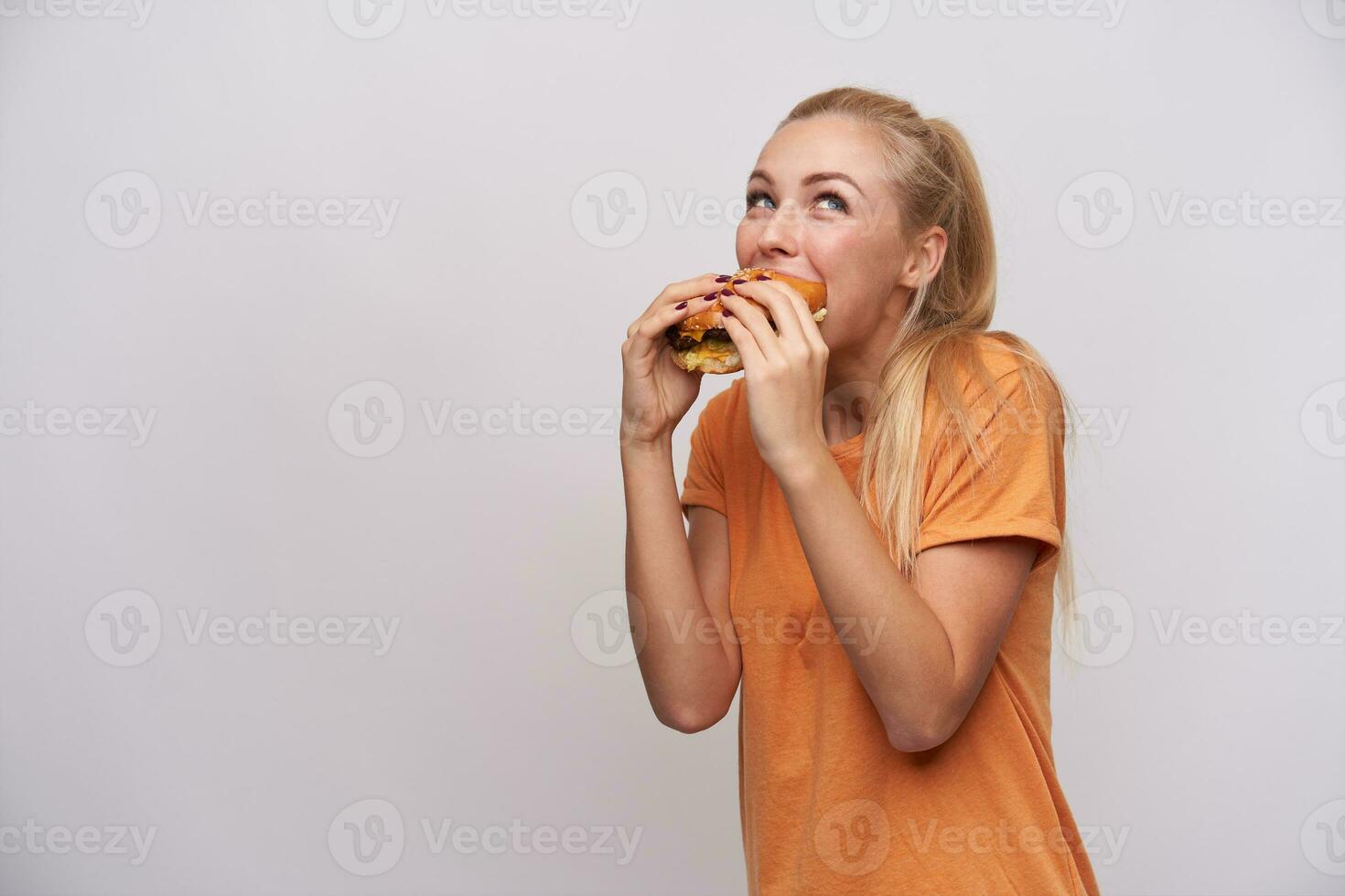 Indoor photo of young cheerful blonde female with casual hairstyle eating hamburger with great pleasure while posing over white background in orange t-shirt