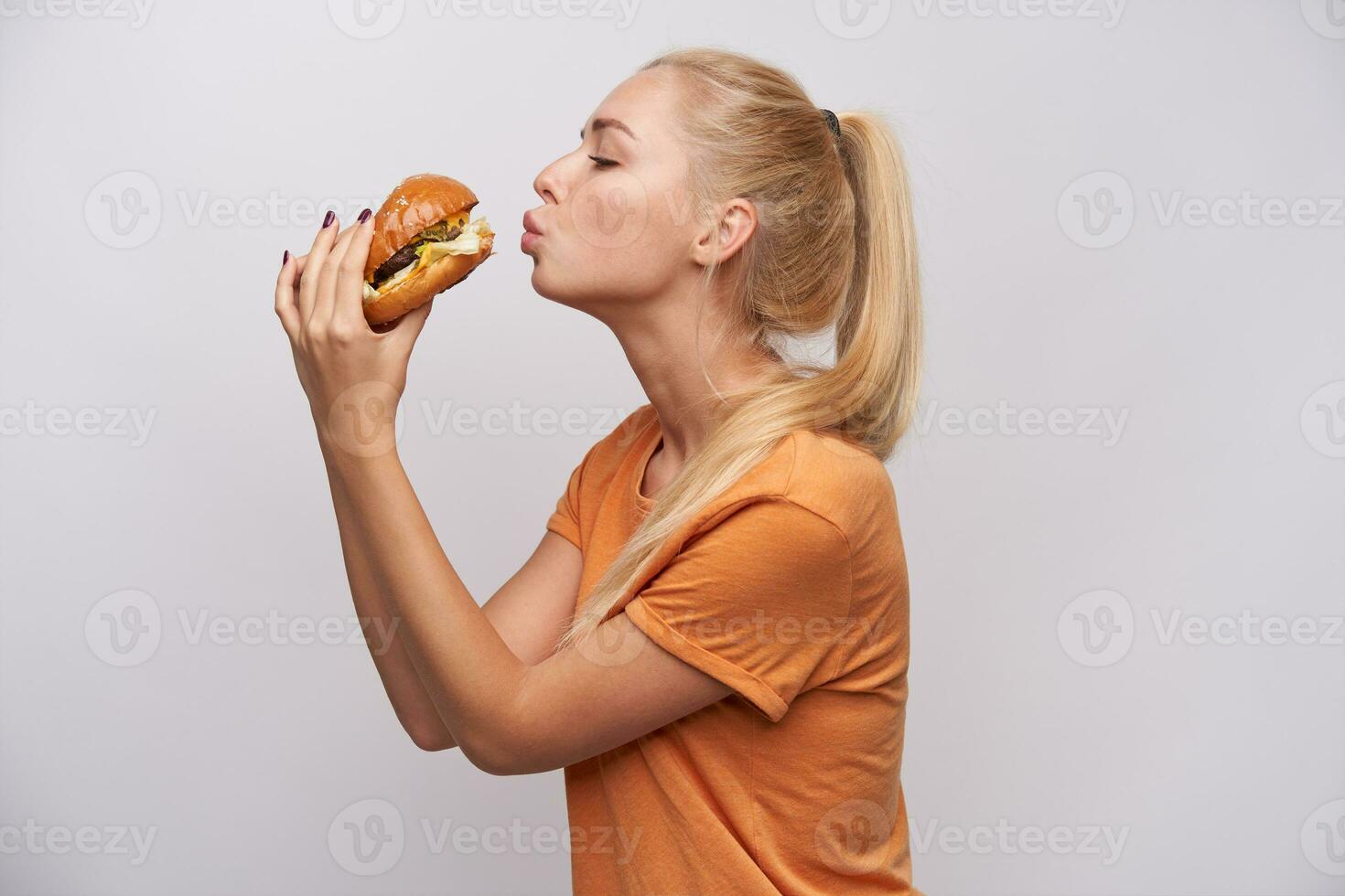 espalda ver de joven largo peludo rubia hembra con cola de caballo peinado levantamiento manos con hambuger y mirando hambriento en él, vestido en casual terminado blanco antecedentes foto