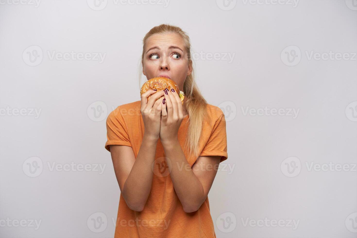 Indoor shot of young pretty afraid blonde lady with ponytail hairstyle eating junk food while nobody seeing her and looking scaredly around, isolated over white background photo