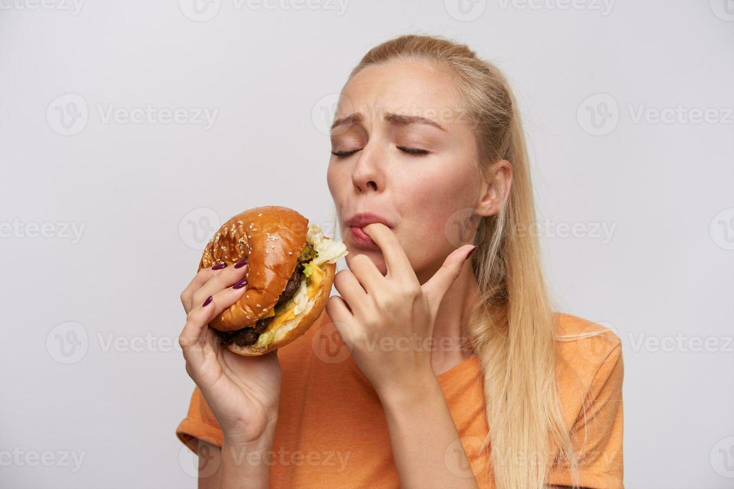 retrato de joven satisfecho rubia hembra con casual peinado saboreo su Fresco hamburguesa con genial Placer y acuerdo ojos cerrado, en pie en contra blanco antecedentes foto