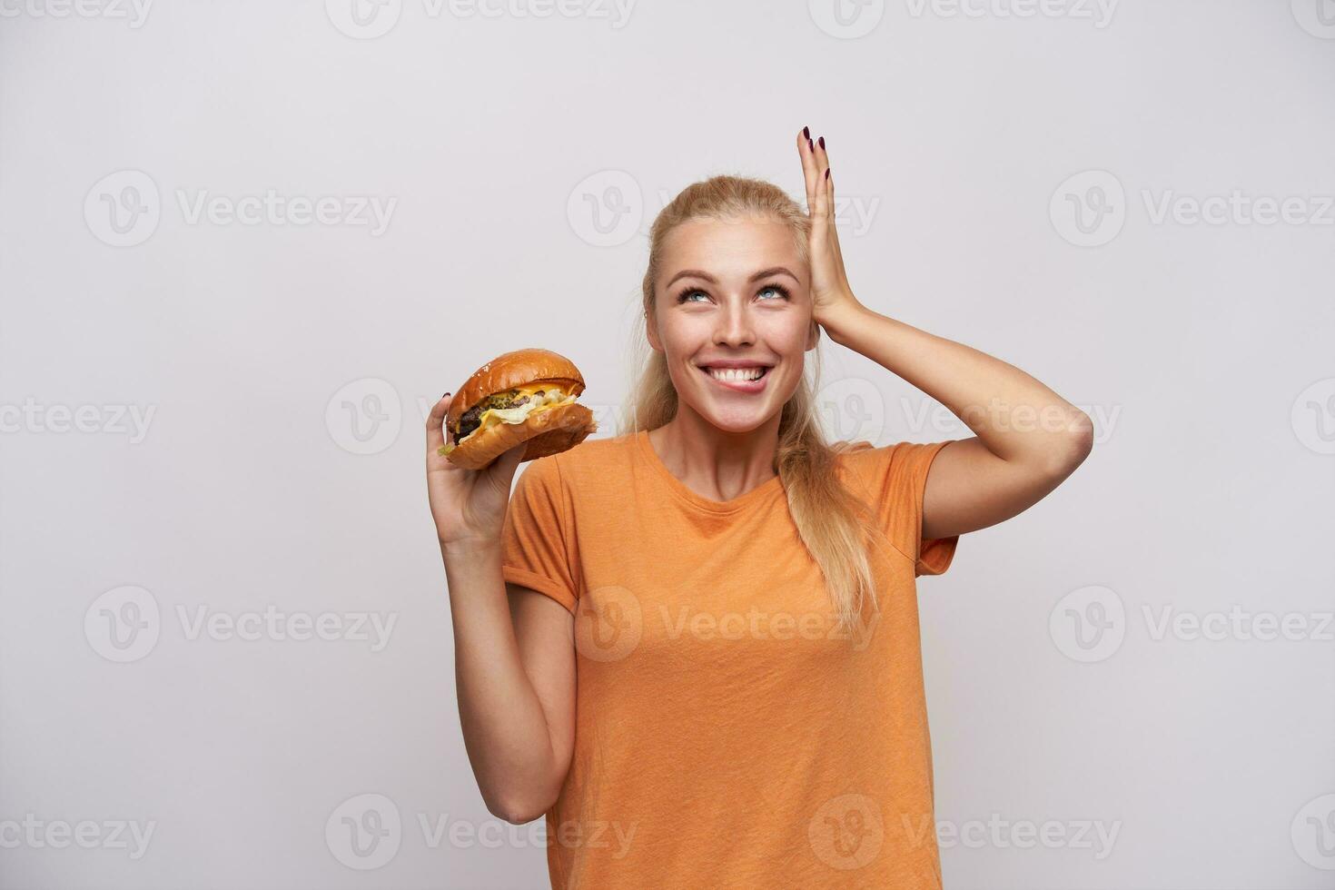 Pleased young long haired blonde lady in orange t-shirt looking upwards cheerfully and foretasting delicious dinner, standing against white background with pleasant smile photo