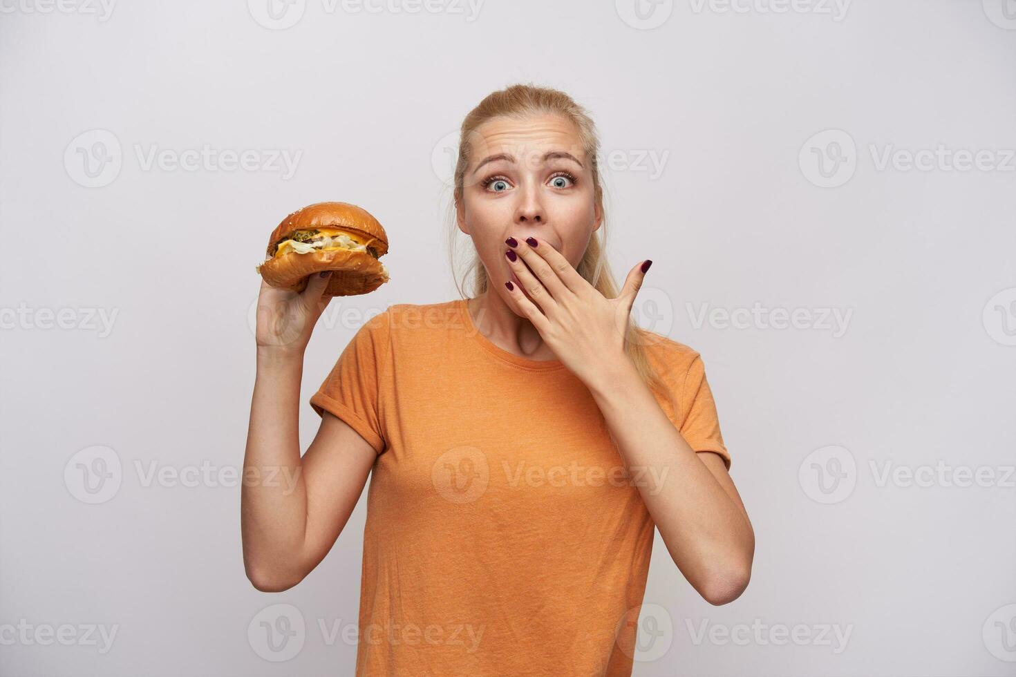 Overjoyed young pretty long haired blonde lady holding delicious burger in raised hand and looking at camera with wide eyes opened, covering her mouth with palm over white background photo