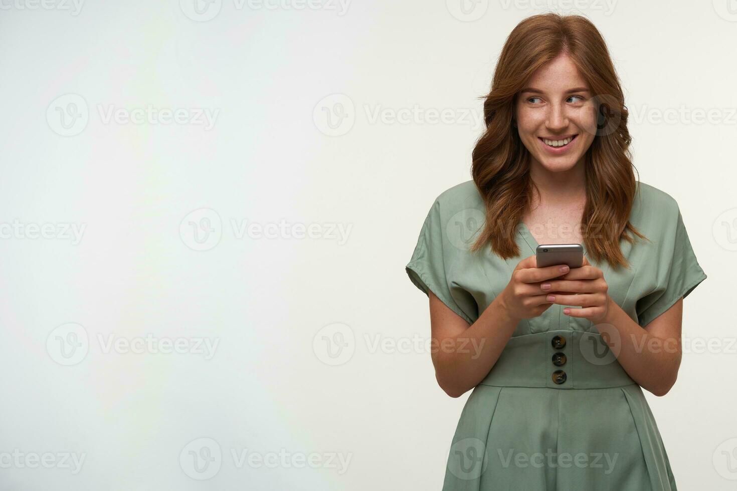 Happy attractive female with red hair posing over white background with mobile phone in hands, looking aside and smiling happily, wearing romantic dress photo