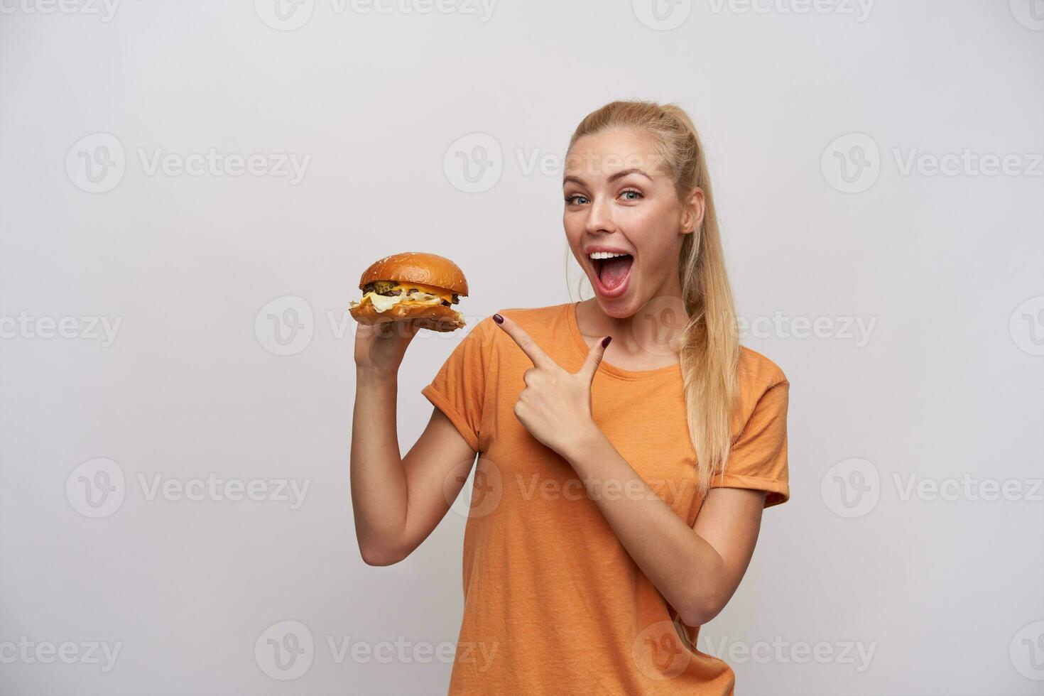 Excited young long haired blonde lady in casual clothes pointing joyfully with forefinger on big fresh burger in raised hand, looking at camera with wide happy smile while posing over white background photo