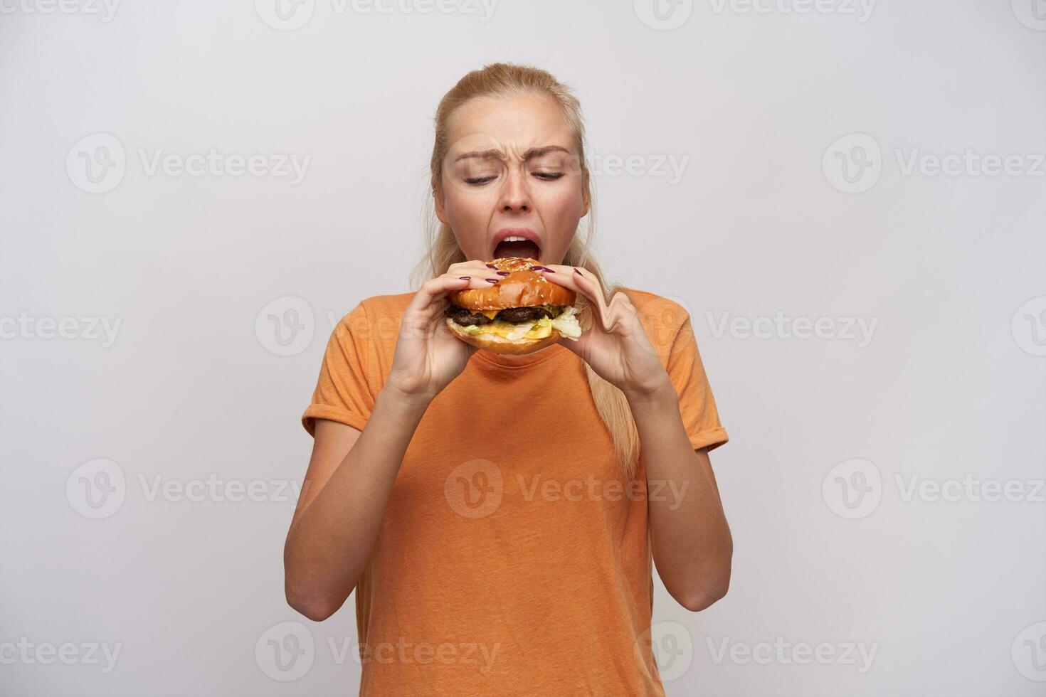 interior foto de joven hambriento largo peludo rubia hembra ceñudo su cara mientras comiendo insaciablemente su sabroso hamburguesa, vestido en naranja camiseta mientras en pie terminado blanco antecedentes