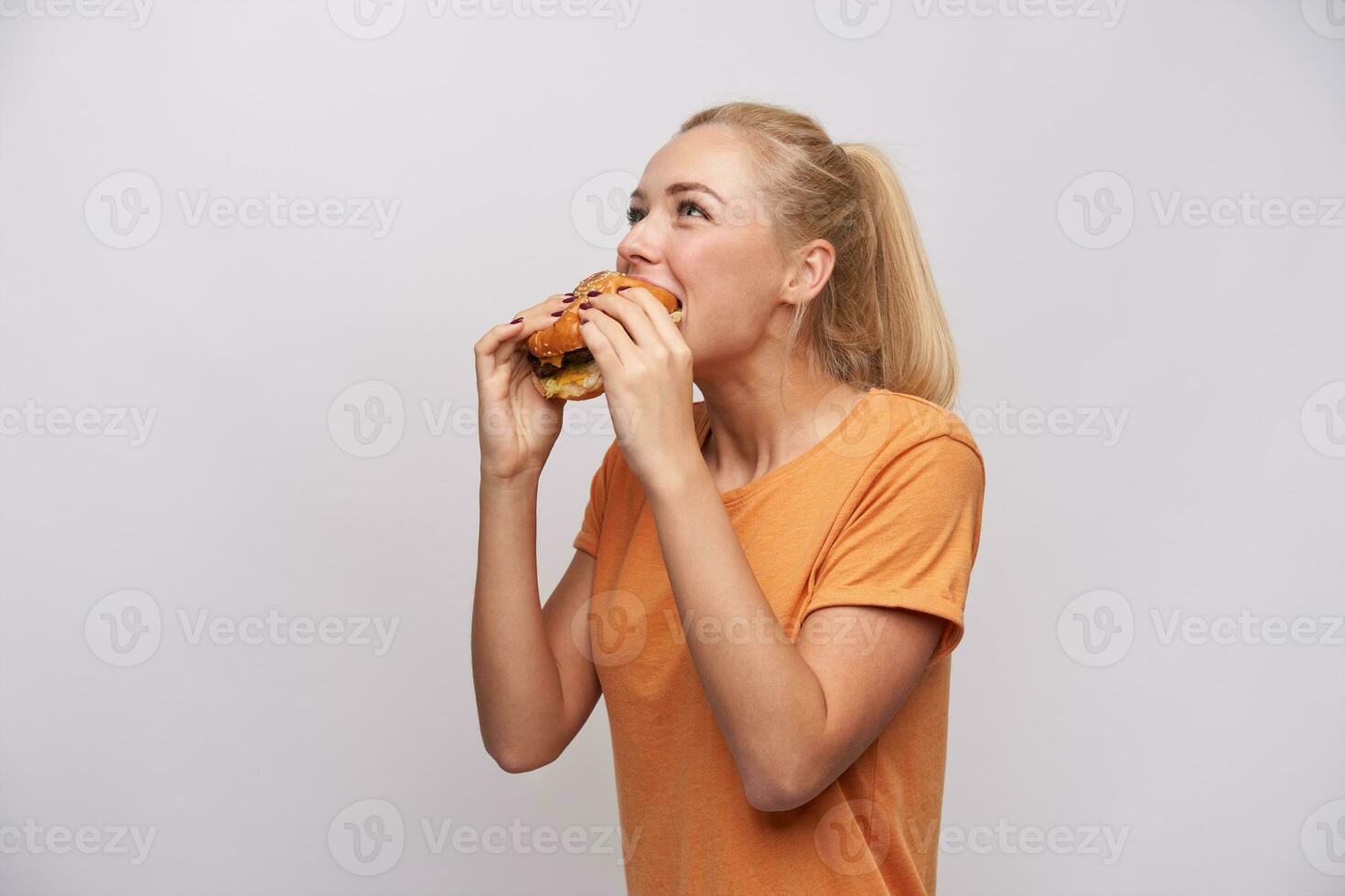Cheerful hungry young blonde female with casual hairstyle biting eagerly off big fresh burger while standing over white background, dressed in orange t-shirt photo