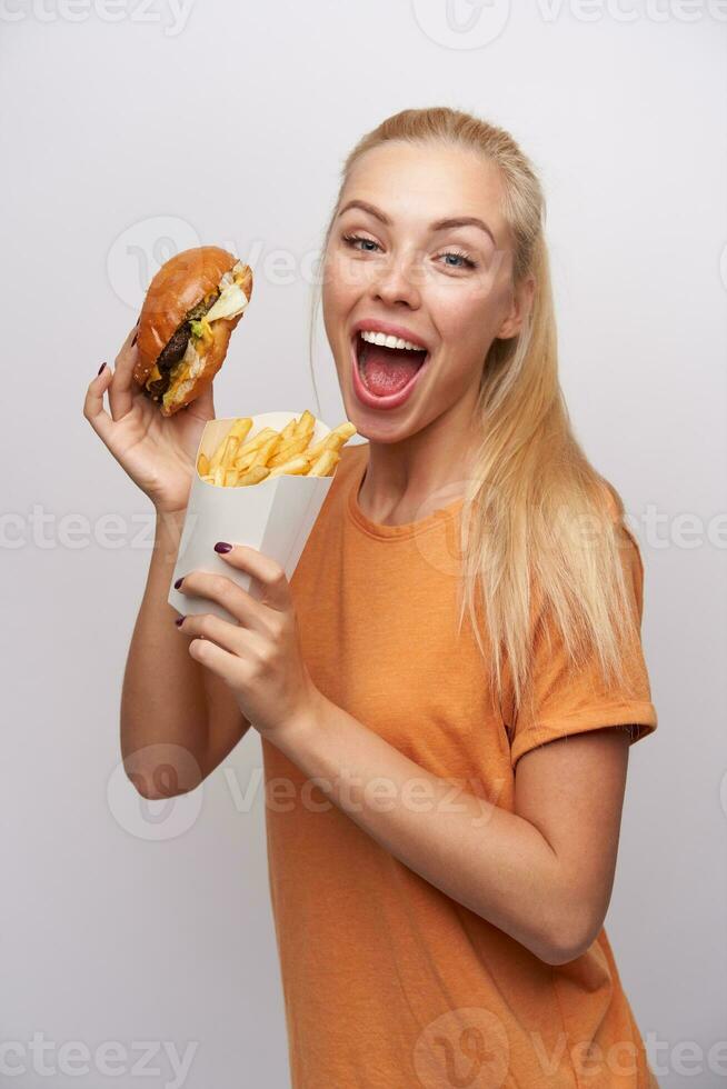 lleno de alegría hermosa joven largo peludo rubia mujer con comida rápida en elevado manos mirando felizmente a cámara con amplio ojos y boca abrió, aislado terminado blanco antecedentes foto