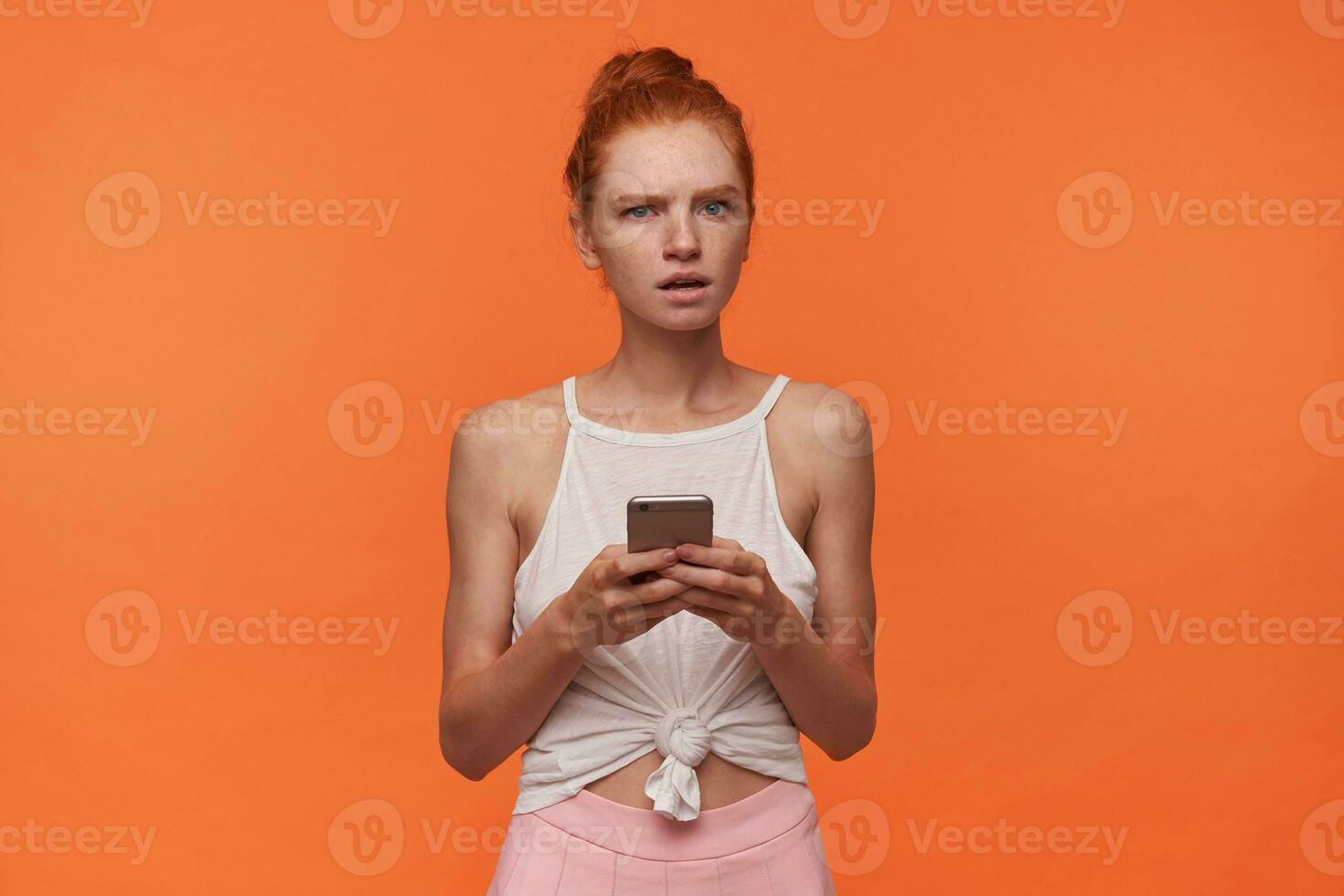 Serious looking young pretty female wearing her red hair in knot, frowning to camera with puzzled face, holding smartphone in hands while posing over orange background in casual clothes photo