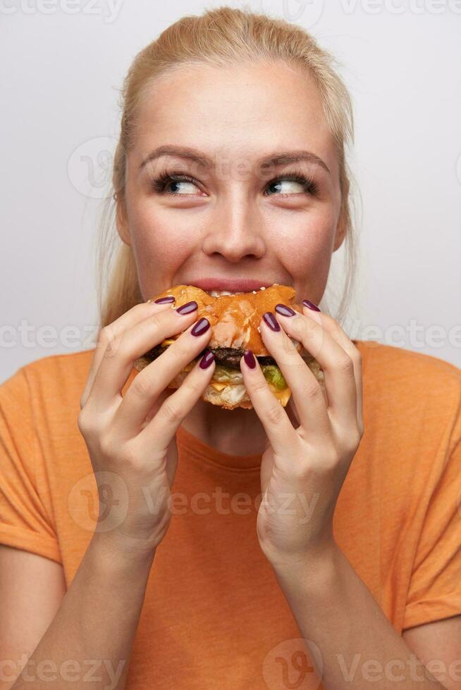 positivo hambriento joven encantador rubia dama con casual peinado mirando alegremente aparte mientras comiendo hamburguesa con genial placer, en pie terminado blanco antecedentes en naranja camiseta foto