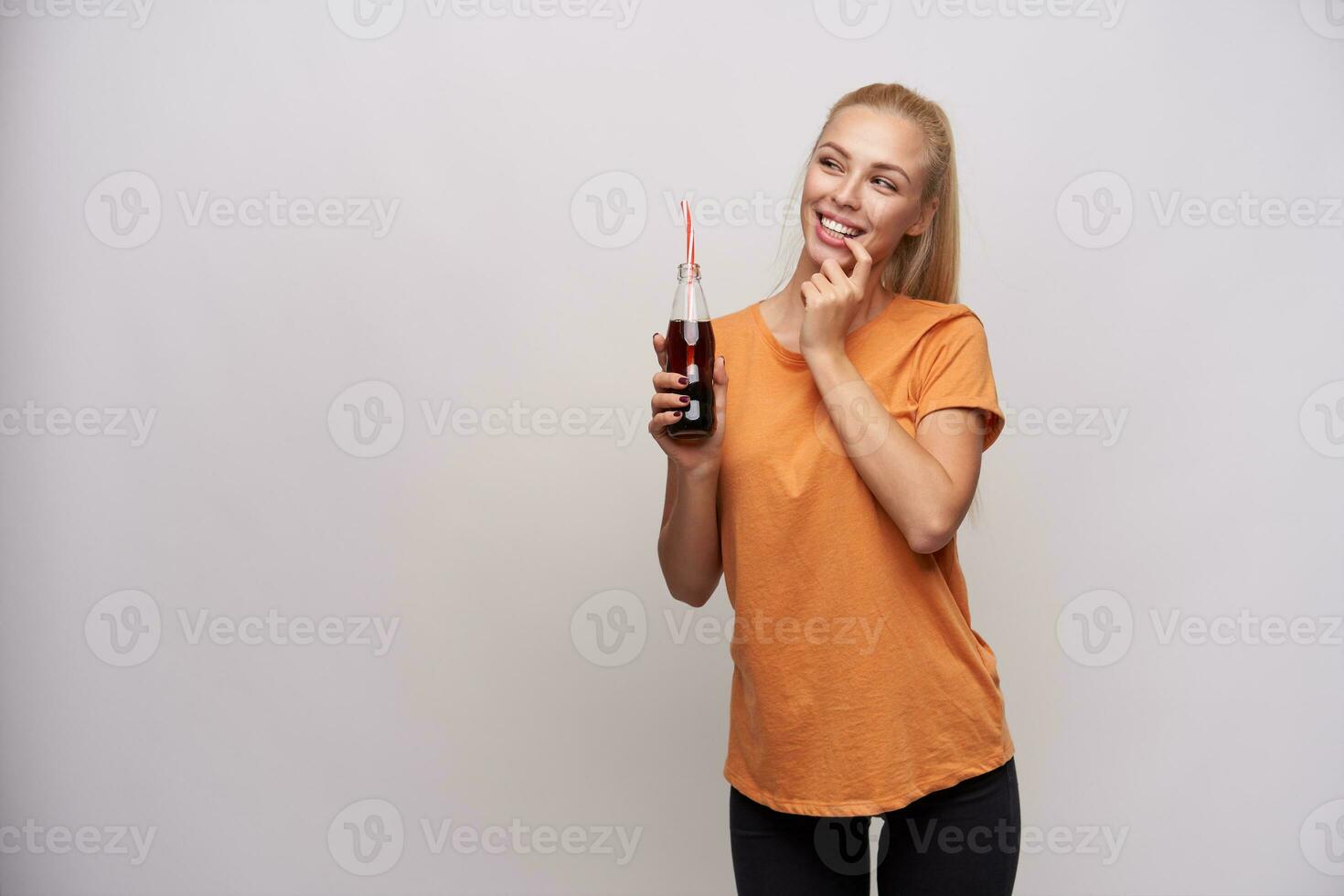 interior foto de alegre joven bonito rubia hembra en casual ropa participación botella de soda en elevado mano y acuerdo dedo índice en debajo del labio mientras mirando aparte con interesar