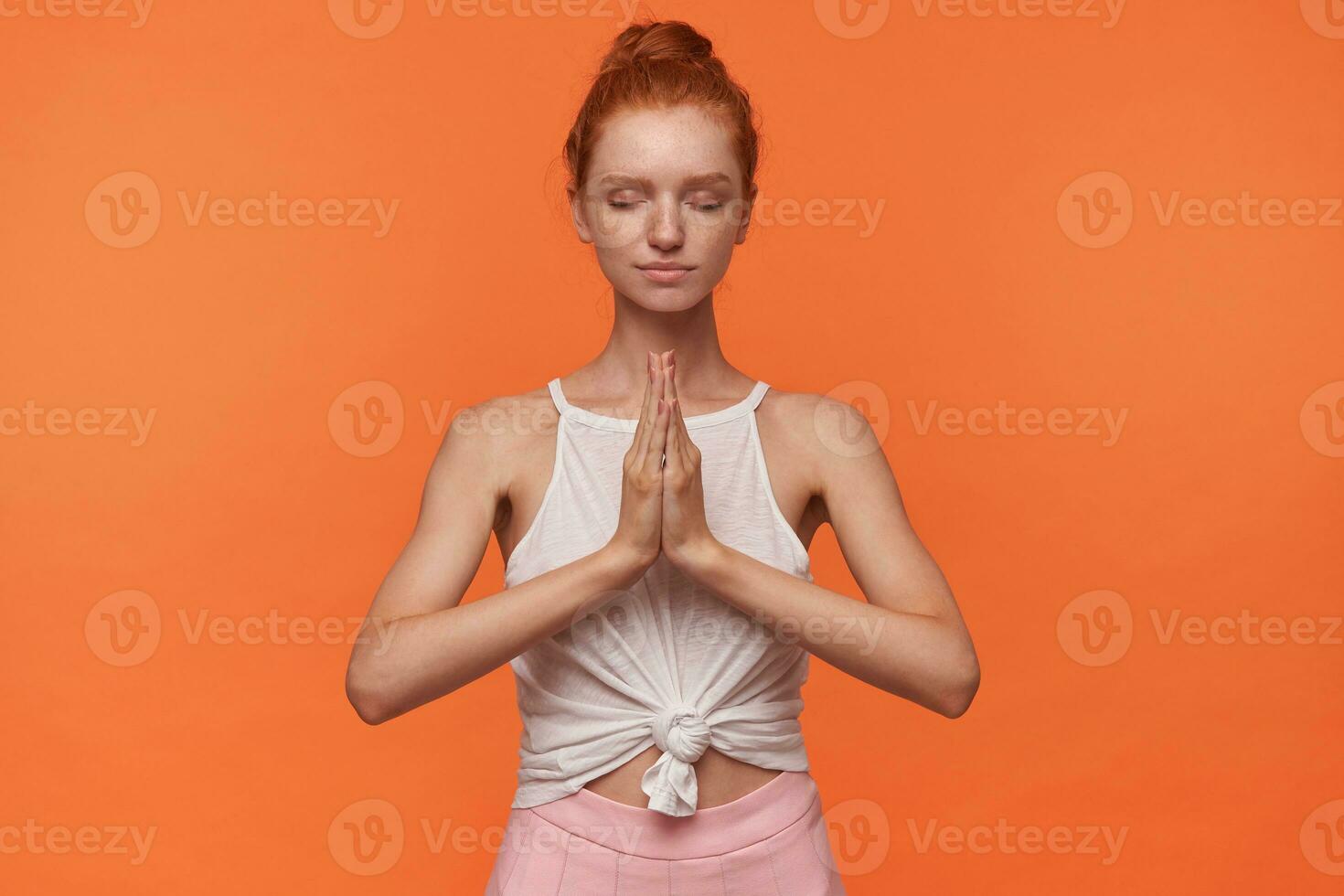 Studio photo of lovely young redhead woman with bun hairstyle wearing white top and pink skirt, raising hands with folded palms in namaste gesture, meditating with closed eyes over orange background