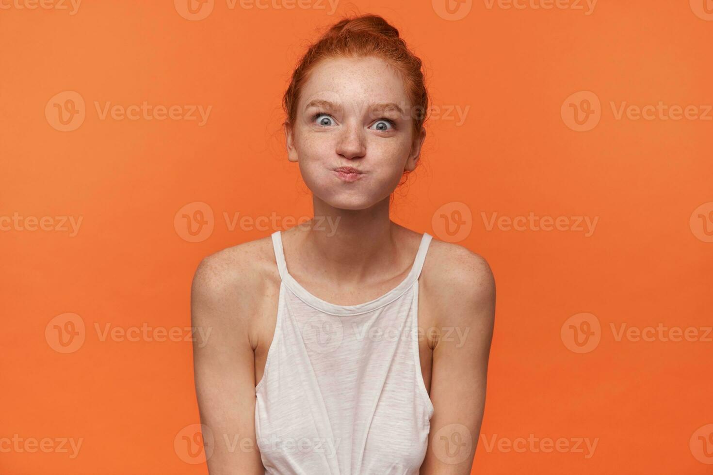 Indoor photo of young funny lady wearing her red hair in knot, looking at camera and puffing out cheeks, fooling and making faces over orange background in white top
