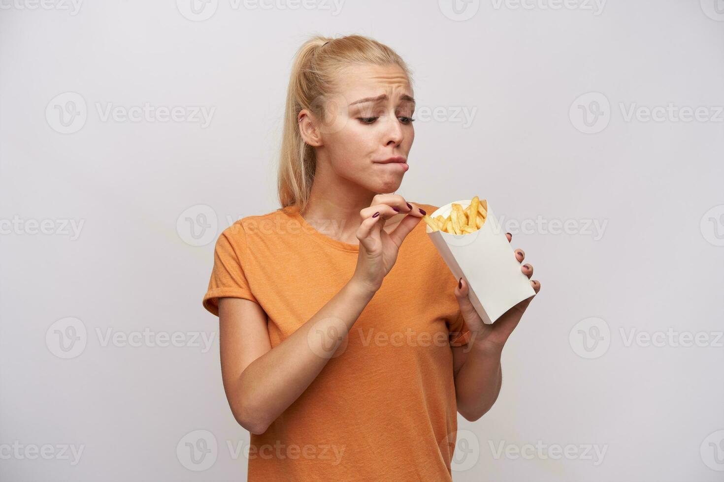 Indoor shot of young long haired blonde lady keeping paper box with french fries and looking excitedly at it, wanting to eat it, but worrying about extra calories, isolated over white background photo