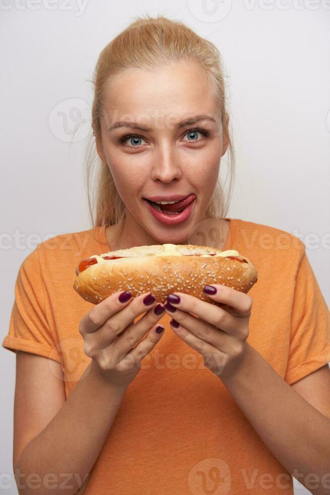 Close-up of hungry blue-eyed excited young blonde lady with tasty hot dog in her hands looking cheerfully at camera and licking her lips, standing against white background photo