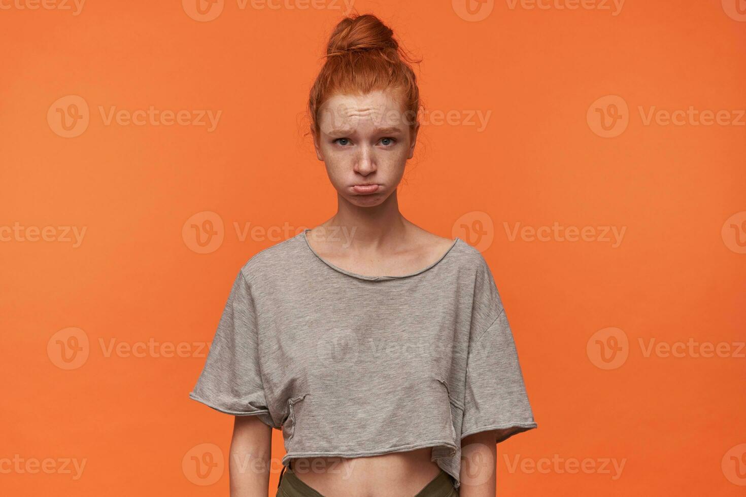 Portrait of cute offended young redhead woman with bun hairstyle wearing casual clothes, posing over orange background with hands down, wrinkling forehead with pursed lips photo