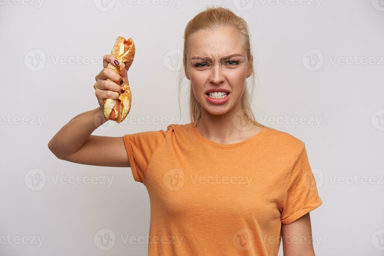 Indoor shot of angry young pretty blonde woman dressed in casual clothes looking violently at camera and crumpling hot dog in raised hand, standing against white background photo
