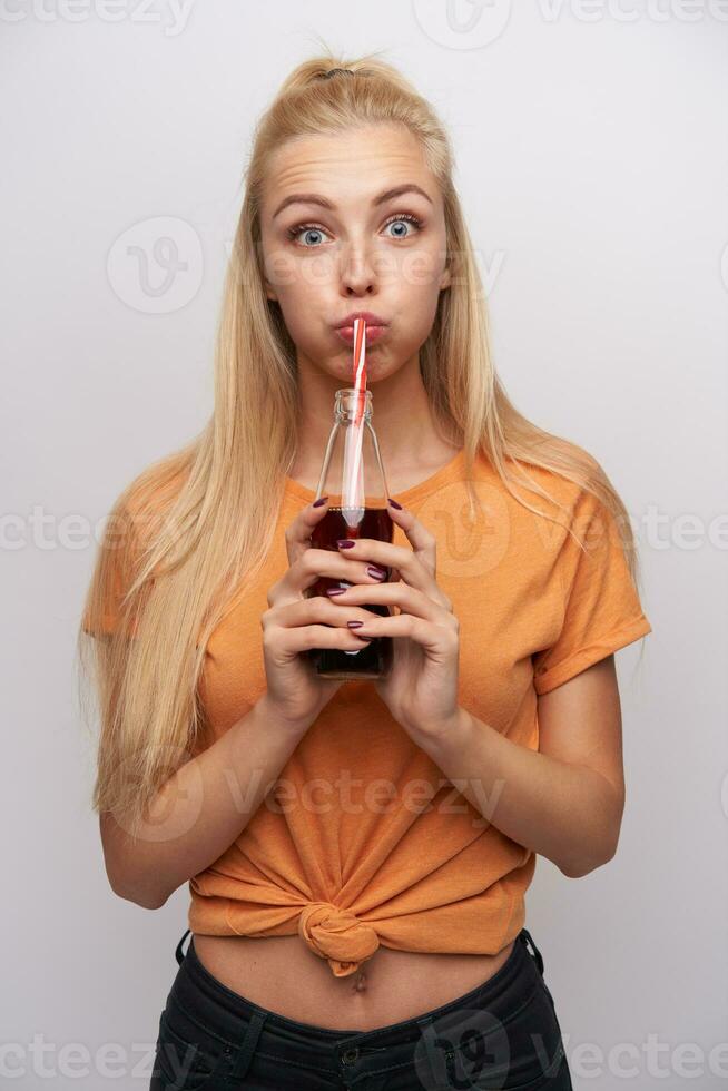 Funny shot of young pretty blue-eyed long haired blonde woman holding glass bottle in hands and fooling while standing over white background, dressed in casual clothes photo