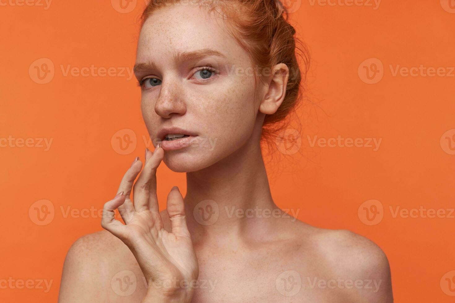 Close-up of romantic young woman with foxy bun hairstyle isolated over orange background, keeping finger on her lips while looking at camera softly, wearing no make-up photo