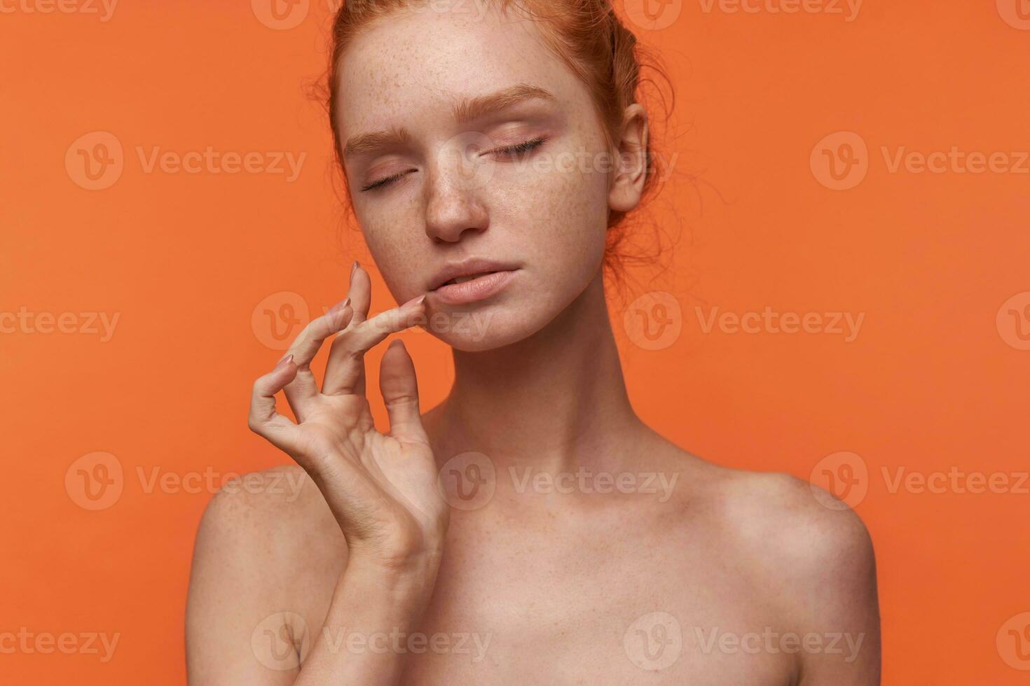 Indoor portrait of young attractive readhead woman wearing hair in knot touching gently her face with hand, standing over orange background with closed eyes photo