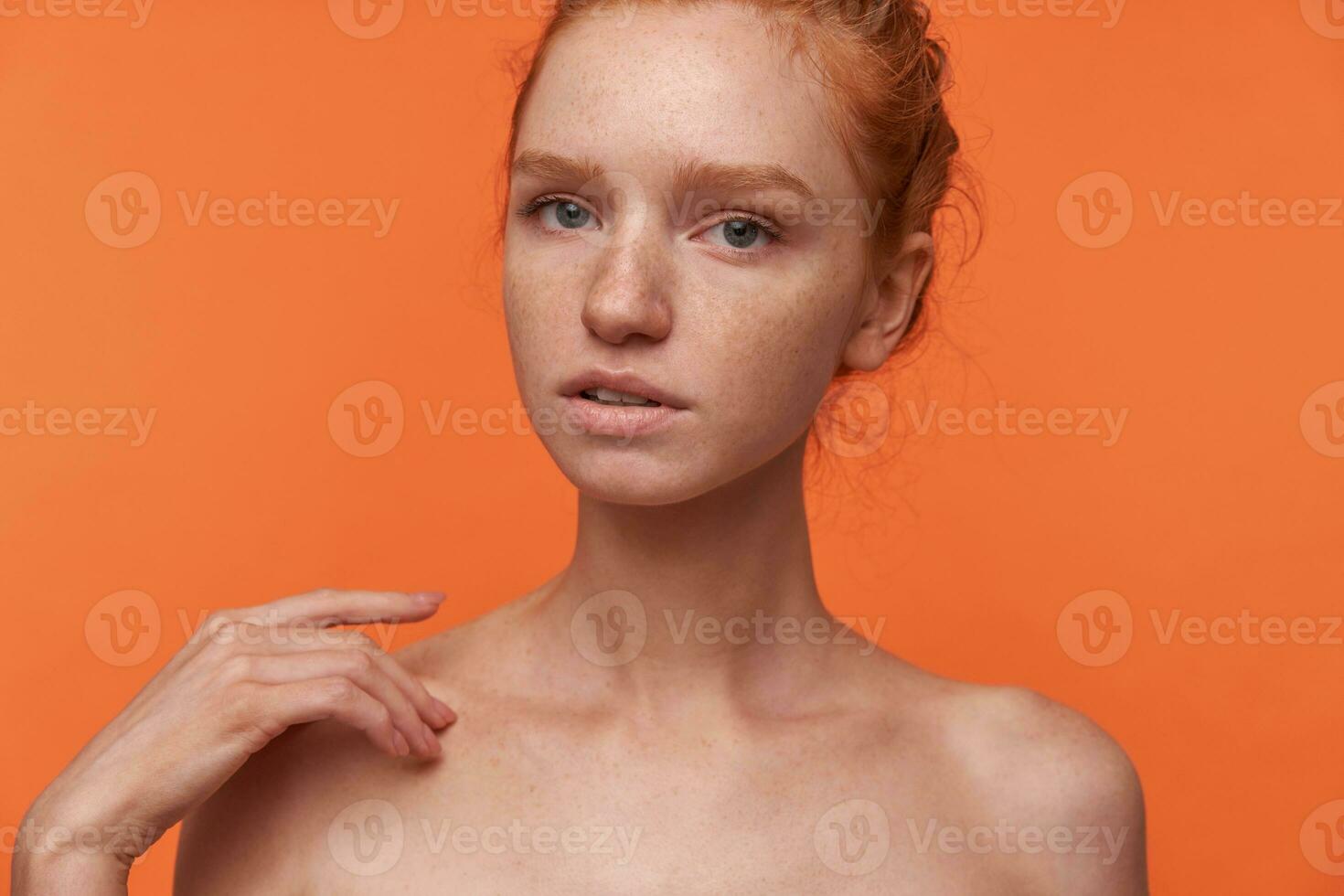 Indoor shot of attractive young female wearing her red hair in bun hairstyle standing over orange background, looking at camera gently with ajar mouth and touching shoulder with raised hand photo