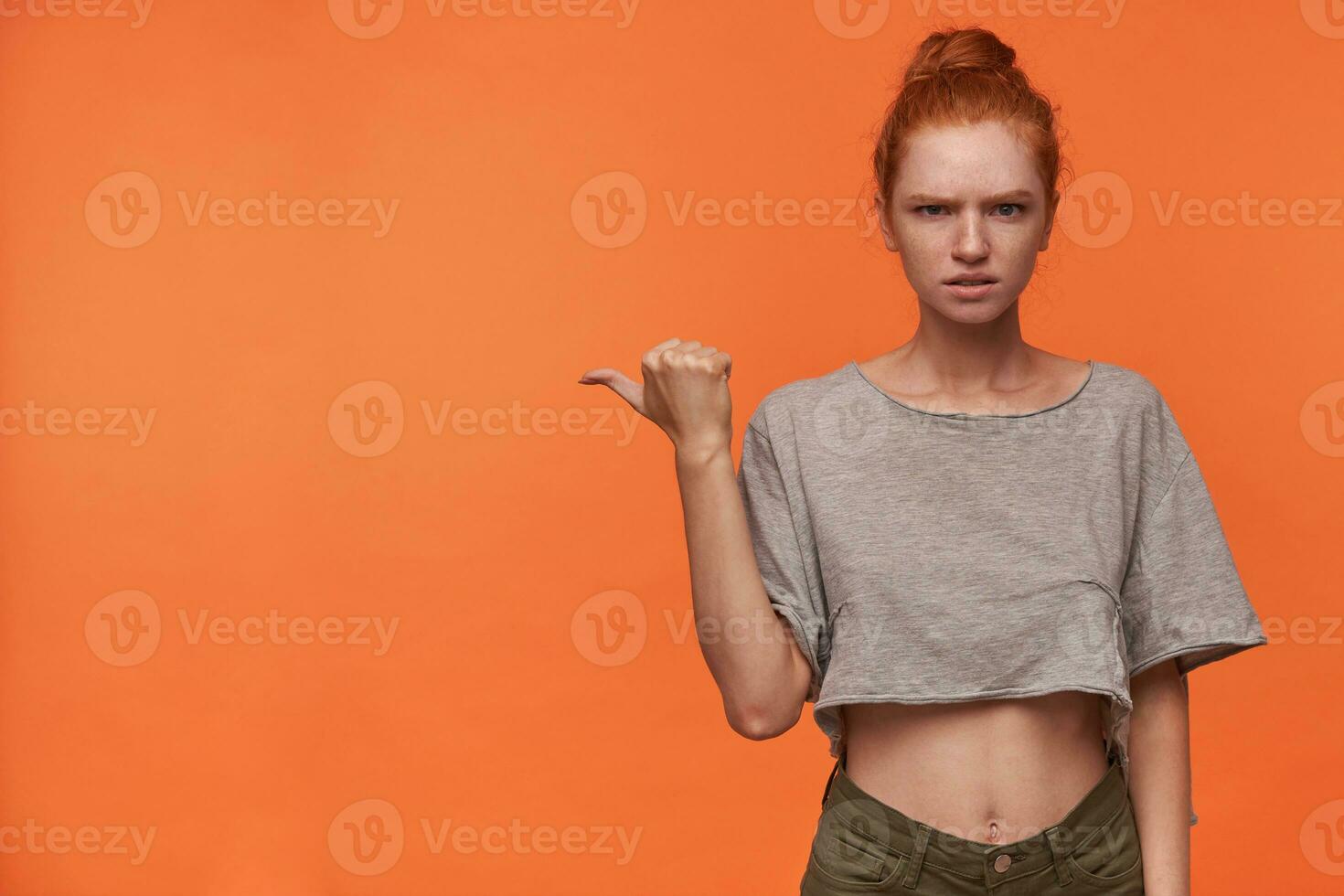 Portrait of serious young readhead woman lwearing her hair in bun, frowning scowlingly to camera and raising hand with thumb pointing aside, standing over orange background photo