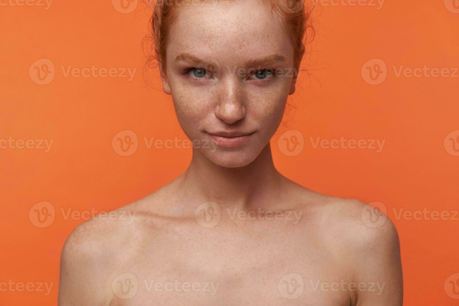 Studio shot of beautiful young lady with red hair in knot standing over orange background with hands down, smiling softly to camera with raised eyebrow. Human emotion facial expression body language photo