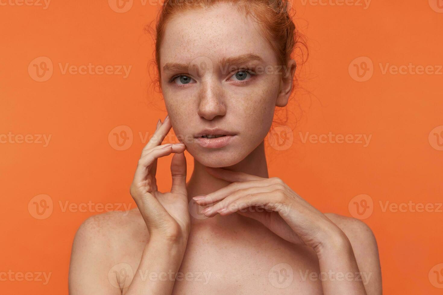 Studio photo of graceful young readhead lady with foxy hair wearing no make-up, raising hand to her face in fine way, touching cheek softly while posing over orange background