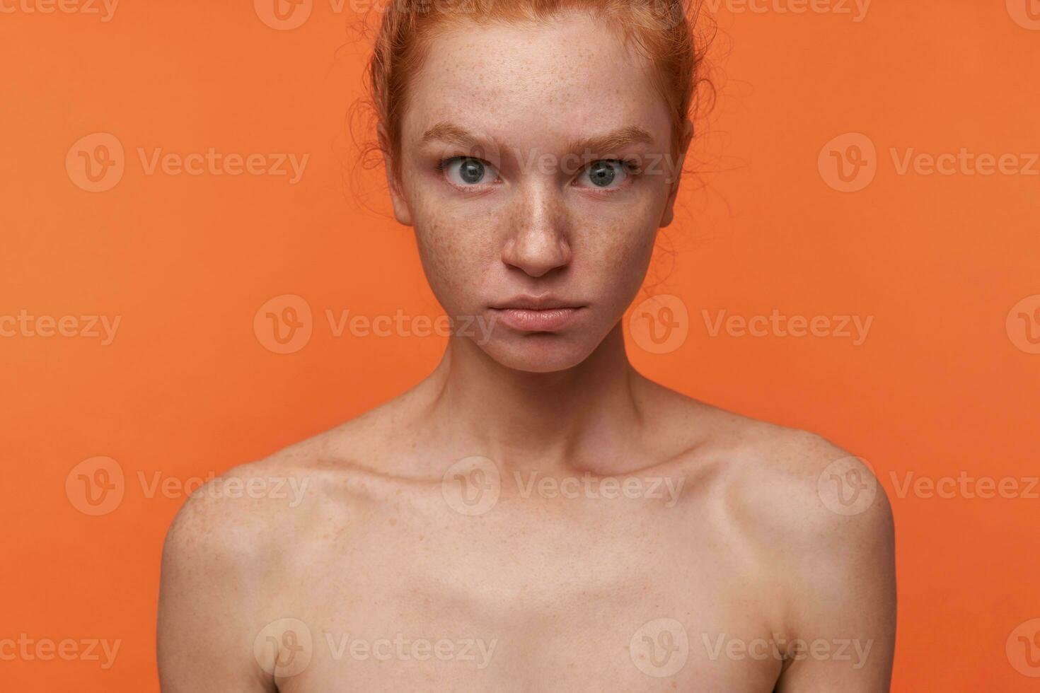 Indoor shot of pretty young readhead female with casual hairstyle and no make-up standing over orange background, looking at camera with puzzled face, rounding eyes amazedly with folded lips photo