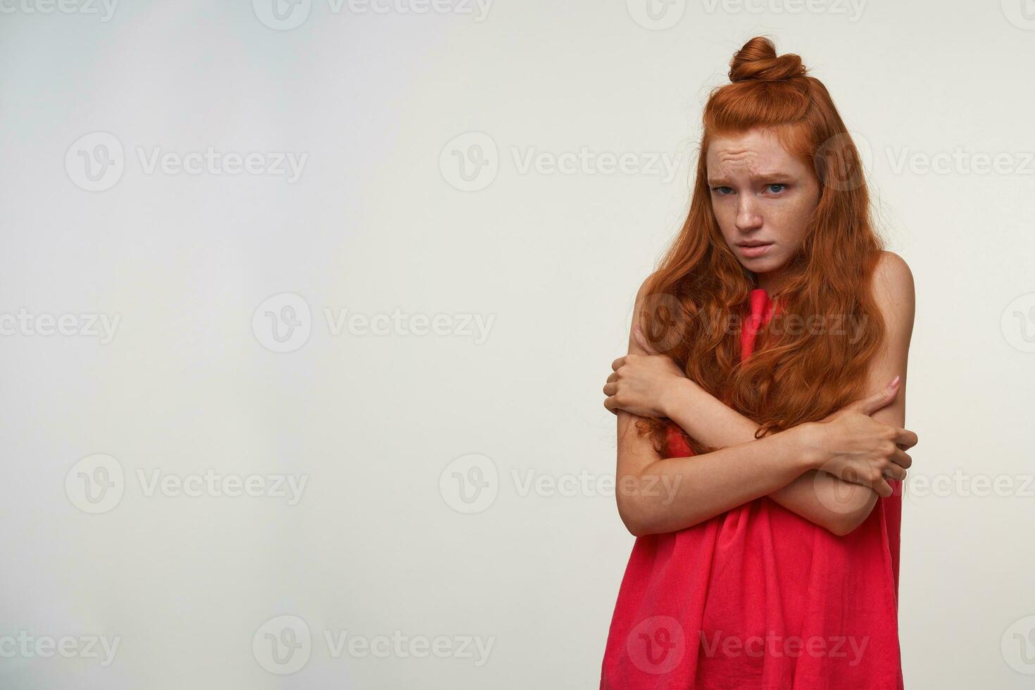 Indoor shot of frightened young readhead female with wavy hair wearing no make up, hugging herself with arms and looking aside with empty eyes, isolated over white background photo