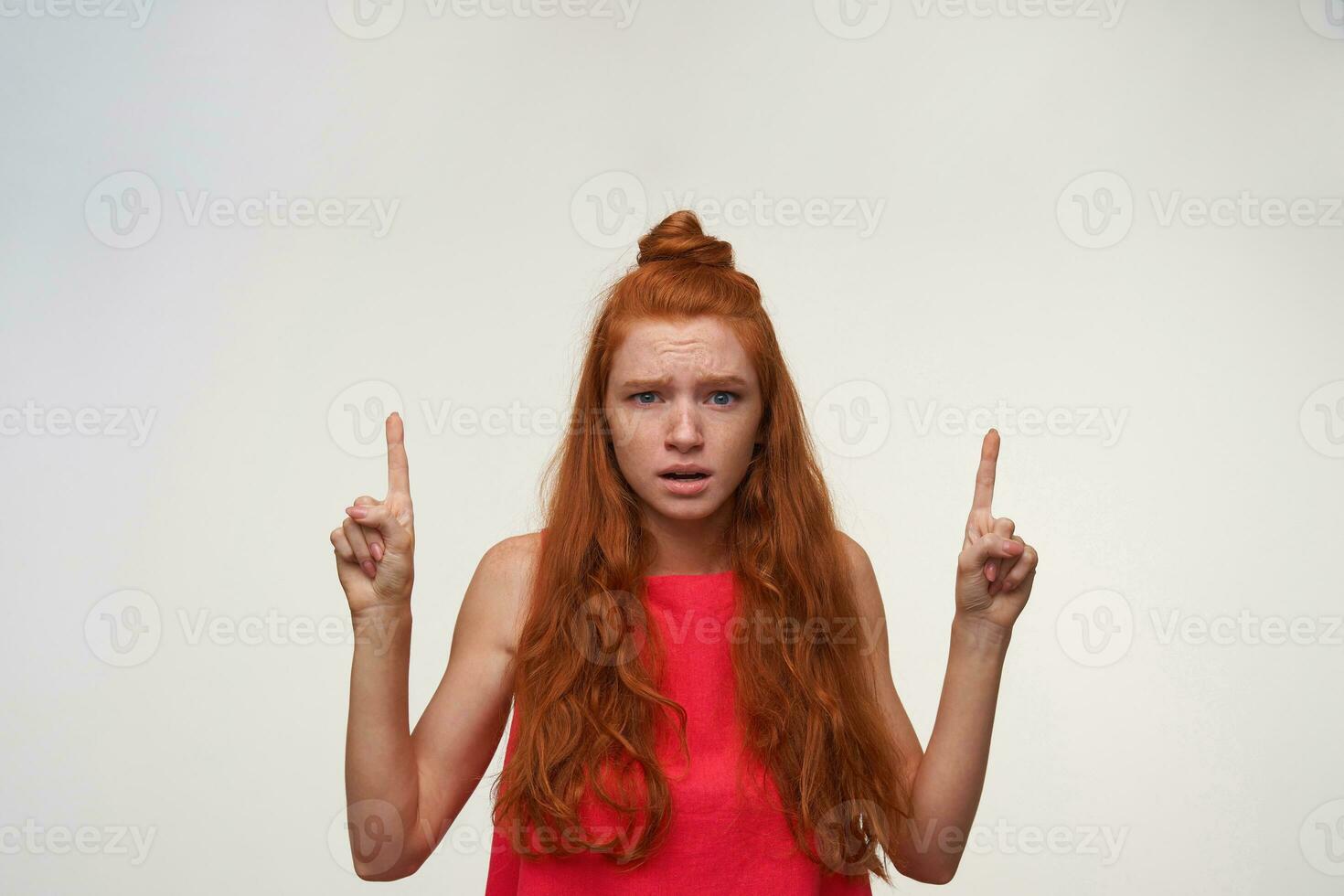 Photo of beautiful young woman with foxy wavy hair wearing no make up posing over white background, looking to camera with confused face and raising forefingers, frowning with wrinkled forehead