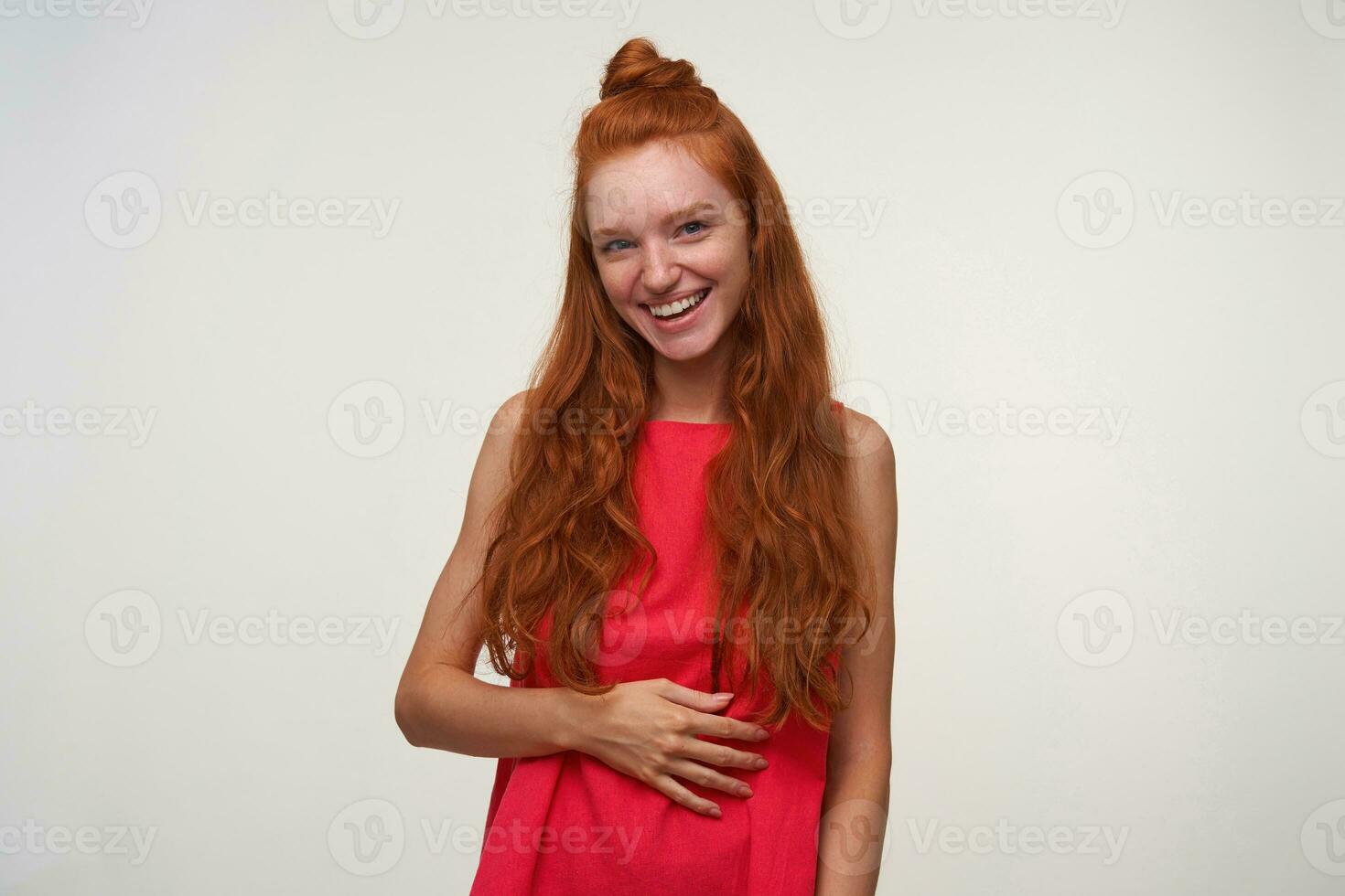 Studio photo of charming young readhead female with wavy hair standing over white background in pink dress, holding palm on her body and smiling positively to camera