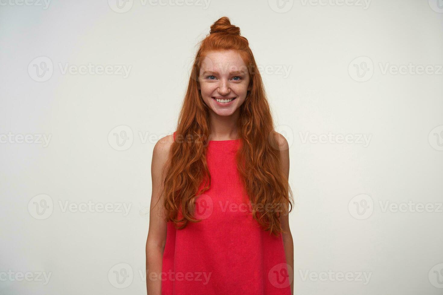 Image of happy lovely young readhead woman with bun posing indoor in pink dress, smiling sincerely to camera over white background with hands down, demonstrating her white perfect teeth photo