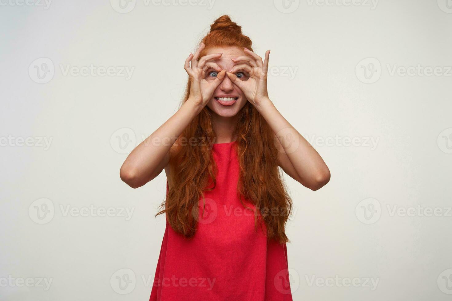 Studio photo of joyful young woman wearing her foxy hair in knot, making ridiculous faces over white background, making eyewear with her hands and showing tongue. Positive emotion facial expression