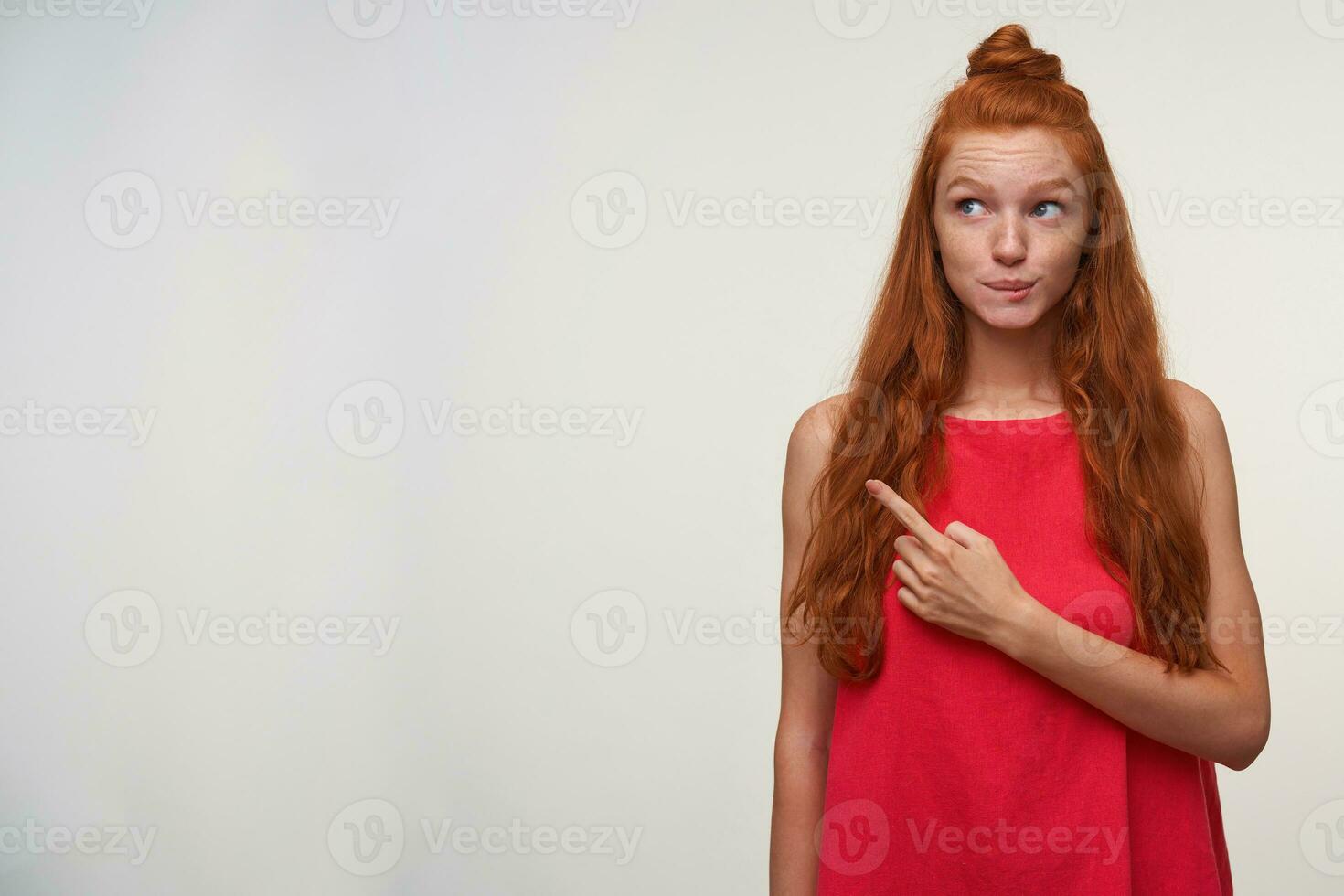 Dreamy lovely young readhead lady with wavy hair wearing no make up, standing over white background in casual dress, showing aside with index finger, raising eyebrows and biting underlip photo