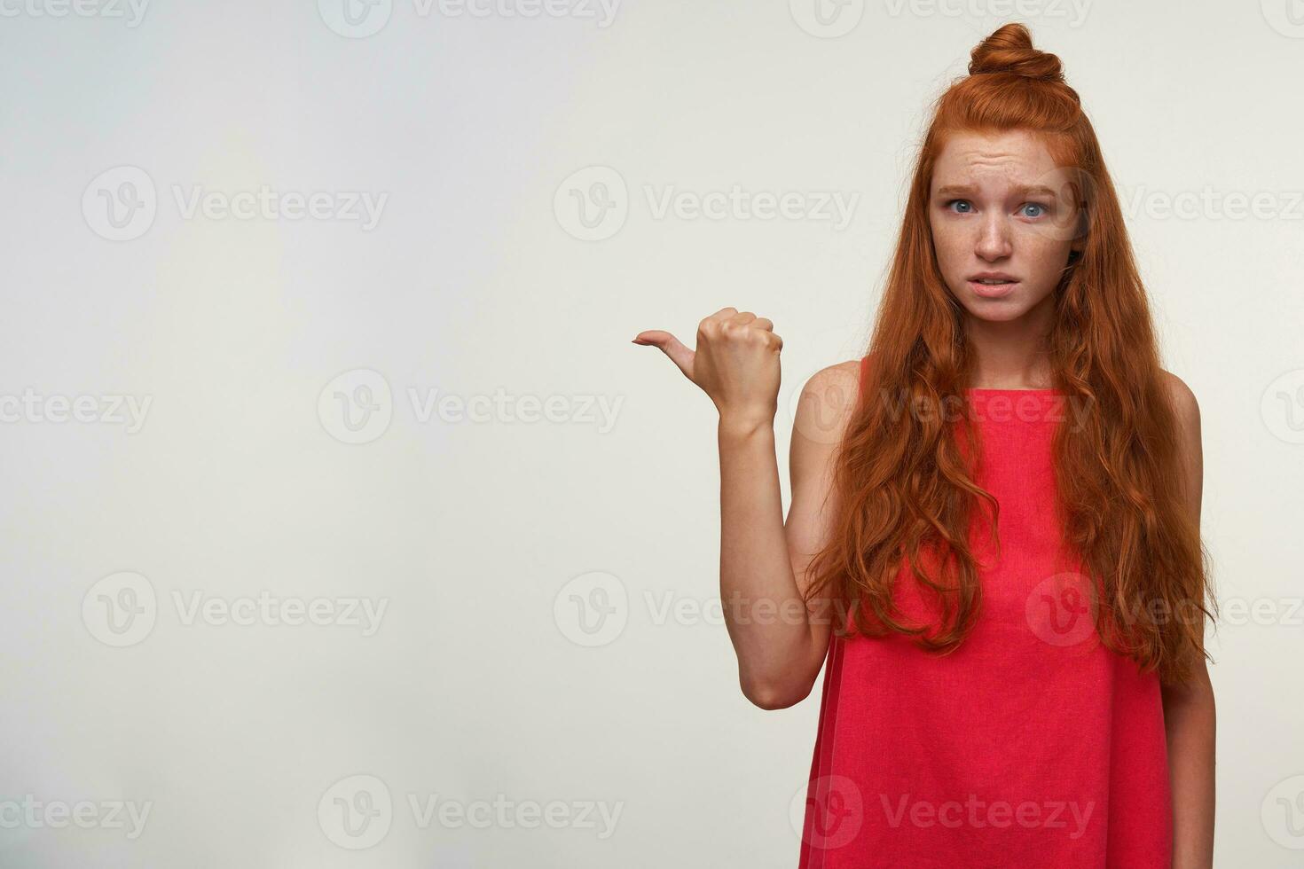 Studio shot of pretty young readhead female with bun hairstyle wearing pink dress, posing over white background with raised thumb and showing away, wrinkling forehead with confused face photo