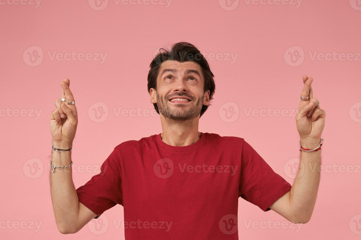 Portrait of young handsome man with trendy haircut standing over pink background in casual clothes, raising hands with crossed fingers for good luck and looking positively upwards photo