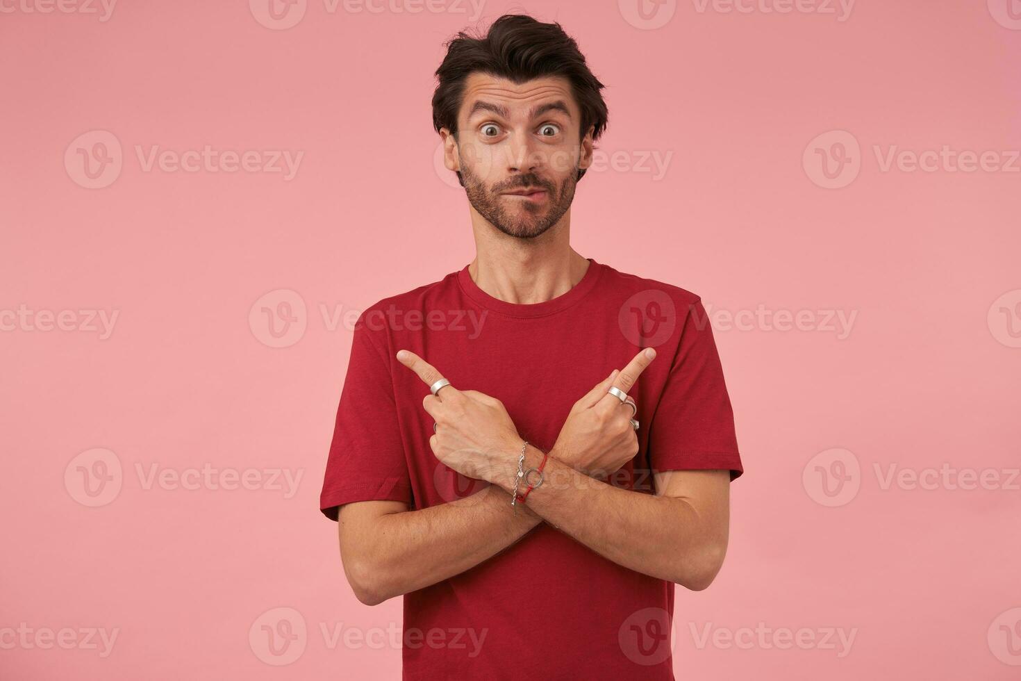 Surprised young man with trendy haircut pointing with forefingers in different directions, standing over pink background in casual clothes, looking at camera with raised eyebrows and biting underlip photo