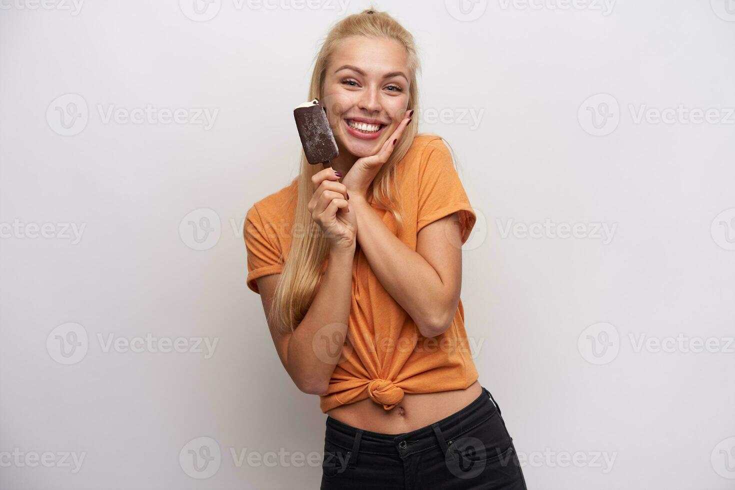 encantador positivo joven rubia dama con casual peinado participación palma en su mejilla y sonriente alegremente a cámara, acuerdo paleta de hielo hielo crema en su mano mientras aislado terminado blanco antecedentes foto