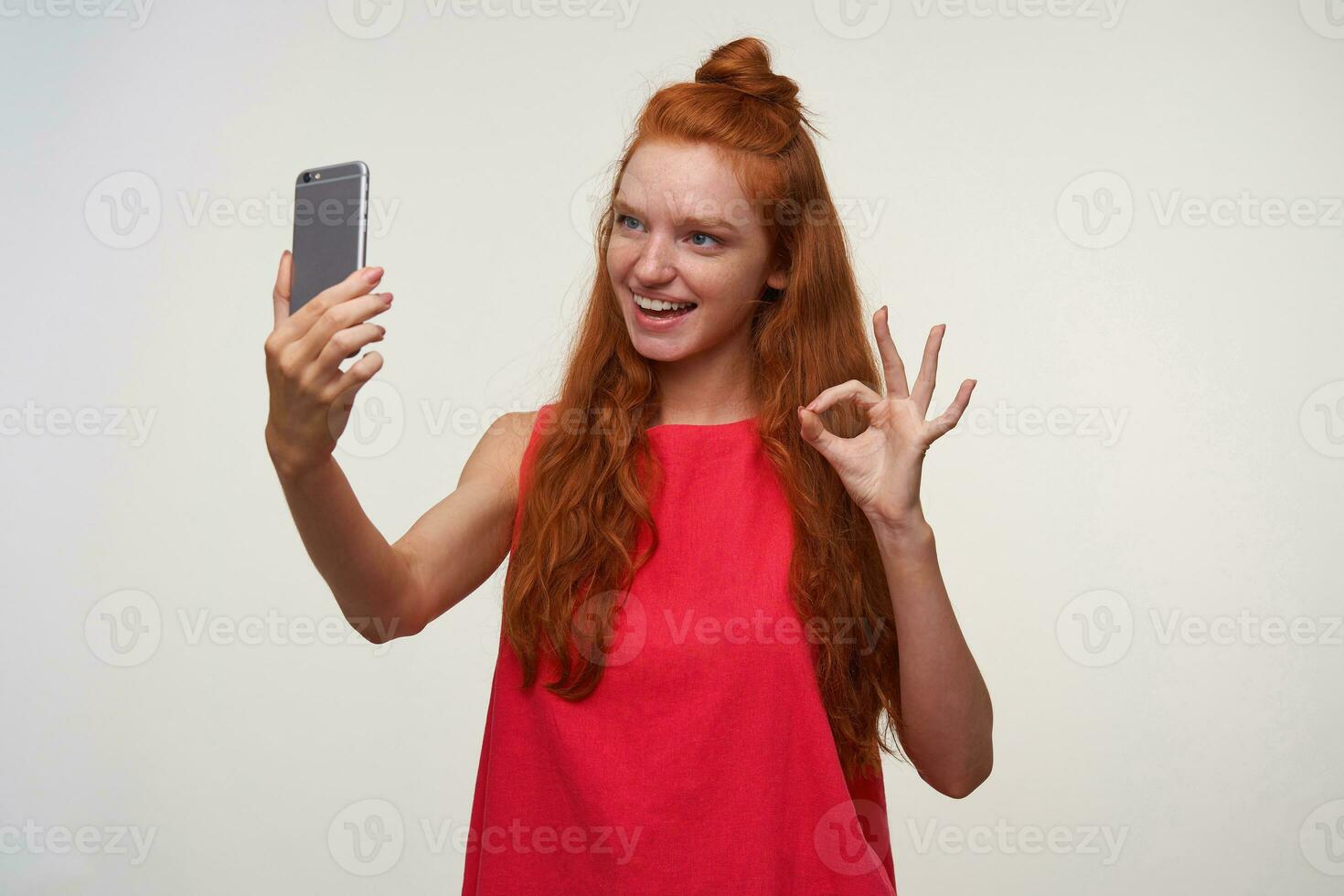retrato de encantador joven dama en rosado vestir vistiendo su astuto pelo en bollo, haciendo foto de ella misma con móvil teléfono, sonriente extensamente a cámara y levantando mano con Okay gesto
