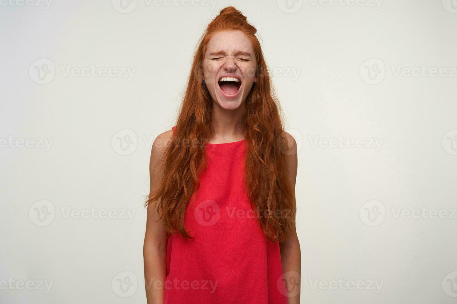 Indoor photo of pretty excited young lady with foxy bun hairstyle wearing pink dress, posing over white background with hands down, shouting loud with closed eyes